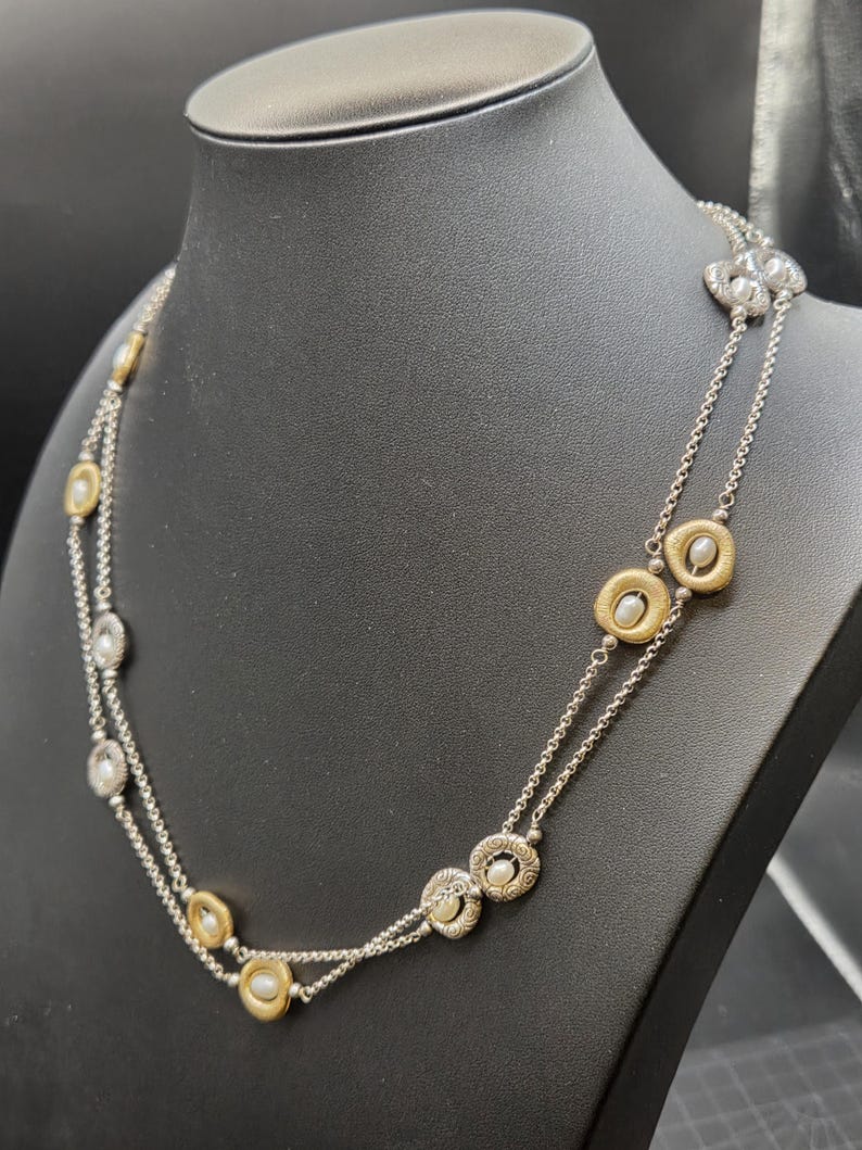 May include: A silver necklace with two strands, featuring gold and silver circular pendants with small, round, white pearls. The necklace is displayed on a black mannequin, highlighting its design and details.