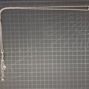 Puede incluir: Collar plateado con un colgante grande en forma de flor con centro y p&eacute;talos color crema. El collar tiene una cadena delicada y un dije m&aacute;s peque&ntilde;o cerca del cierre. El collar se exhibe sobre una superficie gris oscuro.