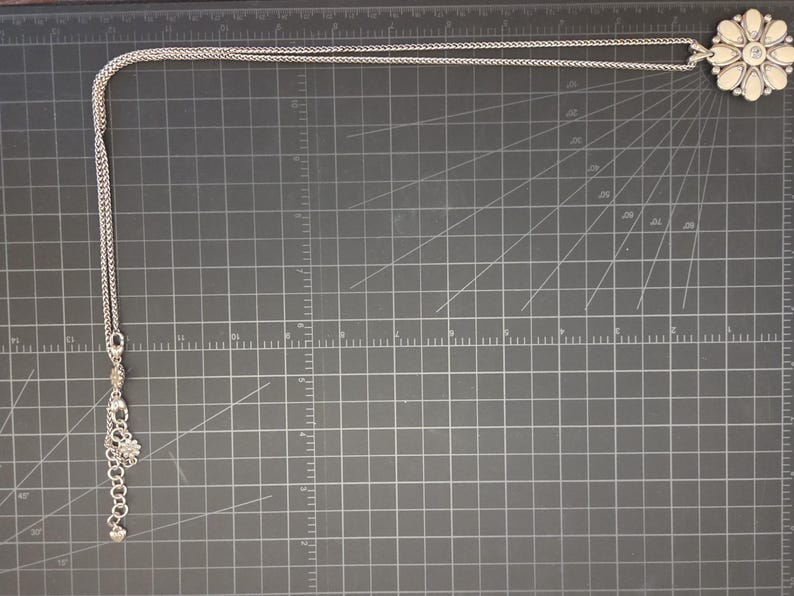 Puede incluir: Collar plateado con un colgante grande en forma de flor con centro y p&eacute;talos color crema. El collar tiene una cadena delicada y un dije m&aacute;s peque&ntilde;o cerca del cierre. El collar se exhibe sobre una superficie gris oscuro.