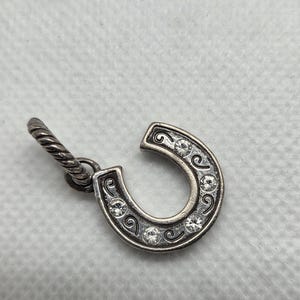 May include: Silver-toned horseshoe charm with clear crystal accents. The horseshoe is detailed with decorative swirls and has a twisted loop for attachment. A small, elegant piece.