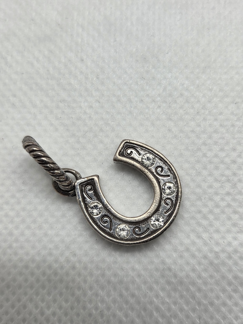 May include: Silver-toned horseshoe charm with clear crystal accents. The horseshoe is detailed with decorative swirls and has a twisted loop for attachment. A small, elegant piece.
