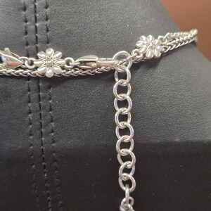 Puede incluir: Collar plateado con un delicado dise&ntilde;o de cadena. El collar presenta peque&ntilde;os dijes en forma de flor y una cadena ajustable con un colgante en forma de coraz&oacute;n. El collar se muestra sobre una superficie negra.