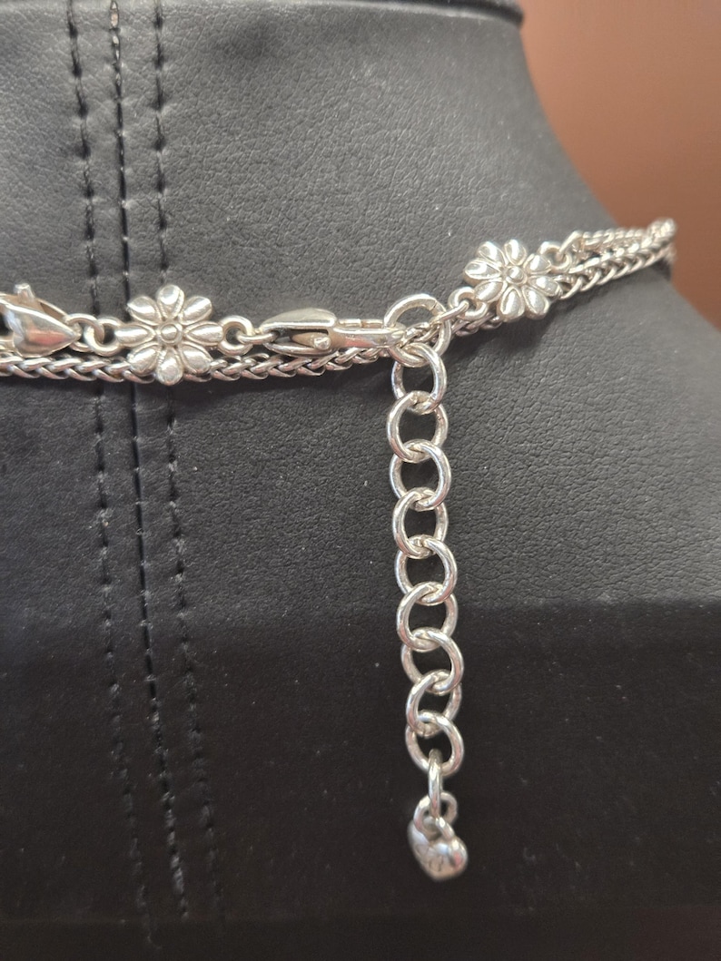 Puede incluir: Collar plateado con un delicado dise&ntilde;o de cadena. El collar presenta peque&ntilde;os dijes en forma de flor y una cadena ajustable con un colgante en forma de coraz&oacute;n. El collar se muestra sobre una superficie negra.