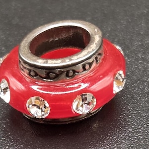 Könnte beinhalten: Eine rote Perle mit silberfarbenem Oberteil und Akzenten. Die Perle hat klare, runde Kristalle und wird wahrscheinlich für die Schmuckherstellung verwendet. Die Perle liegt auf einer dunklen Oberfläche.