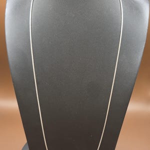 May include: A silver-colored necklace with a delicate chain is displayed on a black mannequin. The chain is thin and appears to be made of metal. The background is a solid brown color.