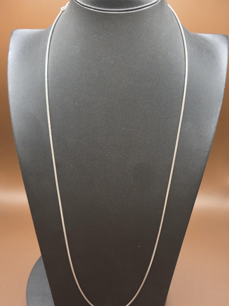 May include: A silver-colored necklace with a delicate chain is displayed on a black mannequin. The chain is thin and appears to be made of metal. The background is a solid brown color.