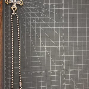 以下が含まれることがあります： ボールチェーンが付いた、銀色のクロス ペンダント ネックレス。クロスには、装飾的な金色のアクセントと透明な宝石が施されています。ネックレスは、グリッド線のある濃い灰色の表面に表示されています。