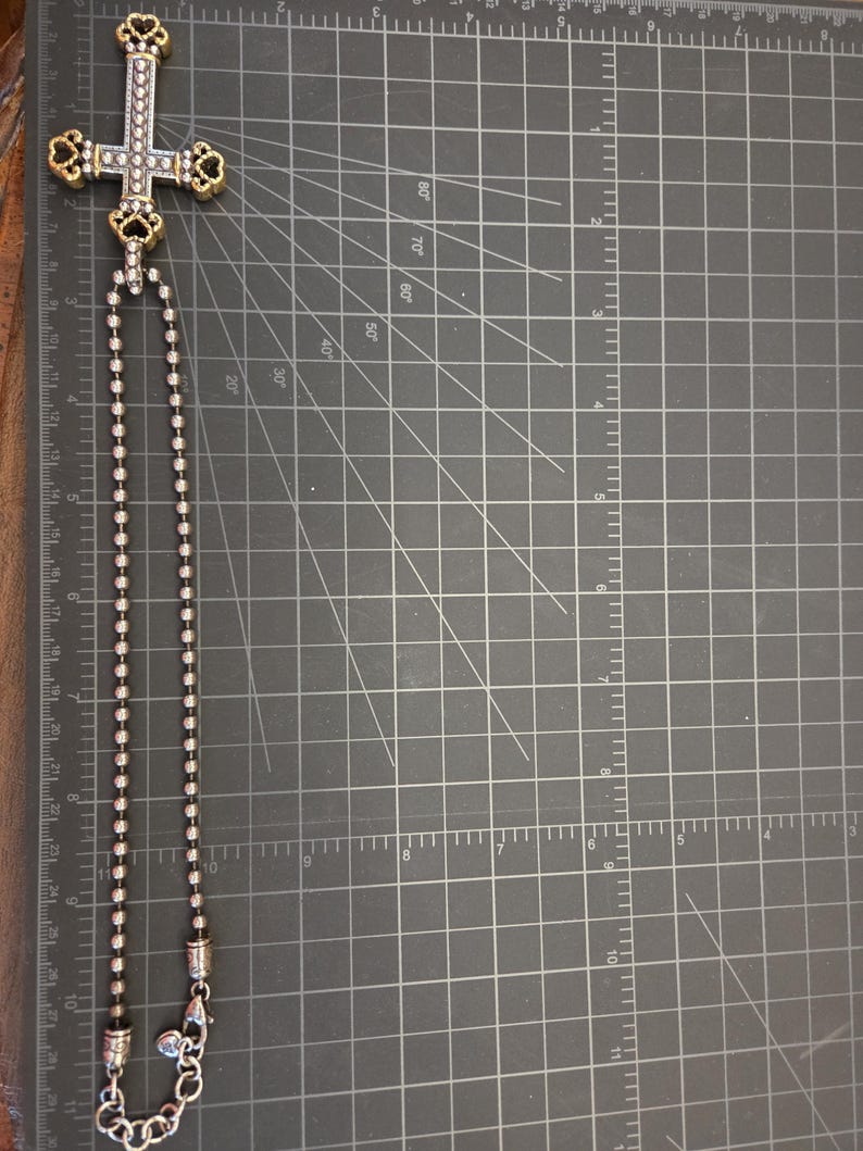 以下が含まれることがあります： ボールチェーンが付いた、銀色のクロス ペンダント ネックレス。クロスには、装飾的な金色のアクセントと透明な宝石が施されています。ネックレスは、グリッド線のある濃い灰色の表面に表示されています。