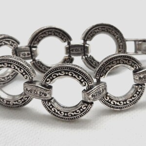 May include: Silver-toned bracelet featuring a series of interconnected, circular links. Each link is adorned with intricate, embossed patterns and designs. The bracelet has a secure clasp closure.