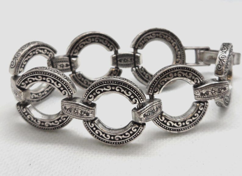 May include: Silver-toned bracelet featuring a series of interconnected, circular links. Each link is adorned with intricate, embossed patterns and designs. The bracelet has a secure clasp closure.