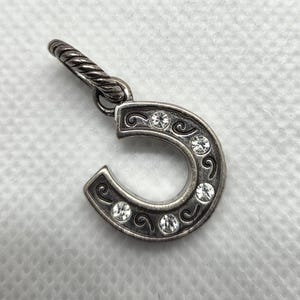 May include: Silver horseshoe charm with clear crystal accents. The horseshoe is detailed with scrollwork and five round crystals. The charm has a twisted bail for attaching to a necklace or bracelet.