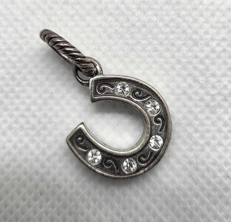 May include: Silver horseshoe charm with clear crystal accents. The horseshoe is detailed with scrollwork and five round crystals. The charm has a twisted bail for attaching to a necklace or bracelet.