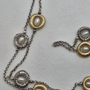 May include: A silver necklace with a double chain design. The necklace features alternating gold-toned and silver-toned oval pendants, each with a faux pearl centre. The gold pendants have a textured finish, while the silver pendants have a scroll design.