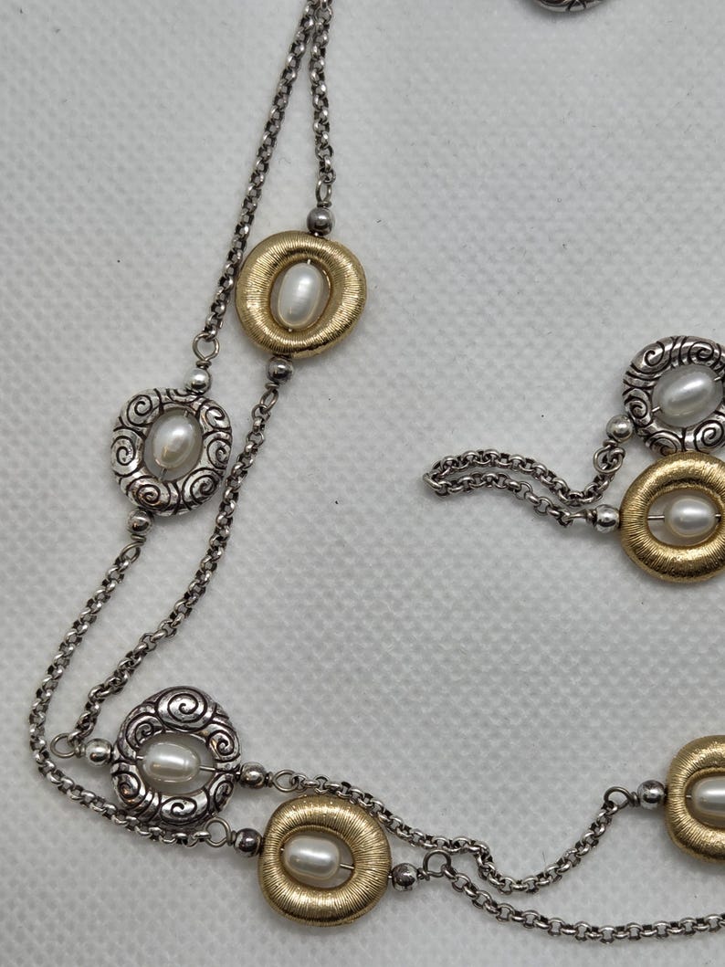May include: A silver necklace with a double chain design. The necklace features alternating gold-toned and silver-toned oval pendants, each with a faux pearl centre. The gold pendants have a textured finish, while the silver pendants have a scroll design.