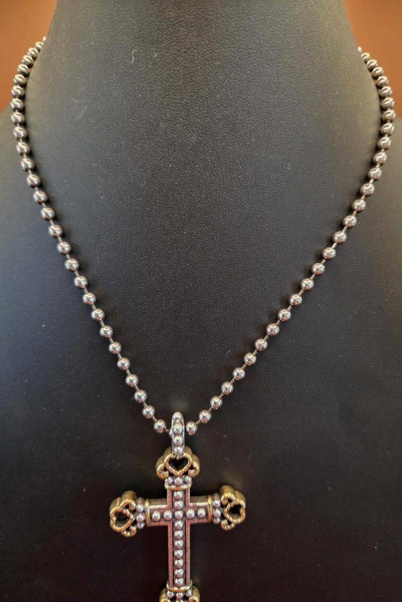 以下が含まれることがあります： 銀色のボールチェーンネックレスと装飾的な十字架ペンダント。十字架は、金色の透かし模様のデザインと、透明なクリスタルのアクセント、中央のクリスタルの列が特徴です。ネックレスは黒い背景に表示されています。