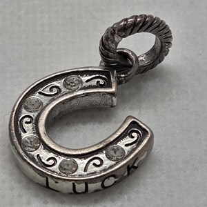 May include: Silver horseshoe charm pendant with clear crystal accents and the word "LUCK" engraved on the bottom. The horseshoe has decorative swirls and a twisted loop for attaching to a chain or bracelet.