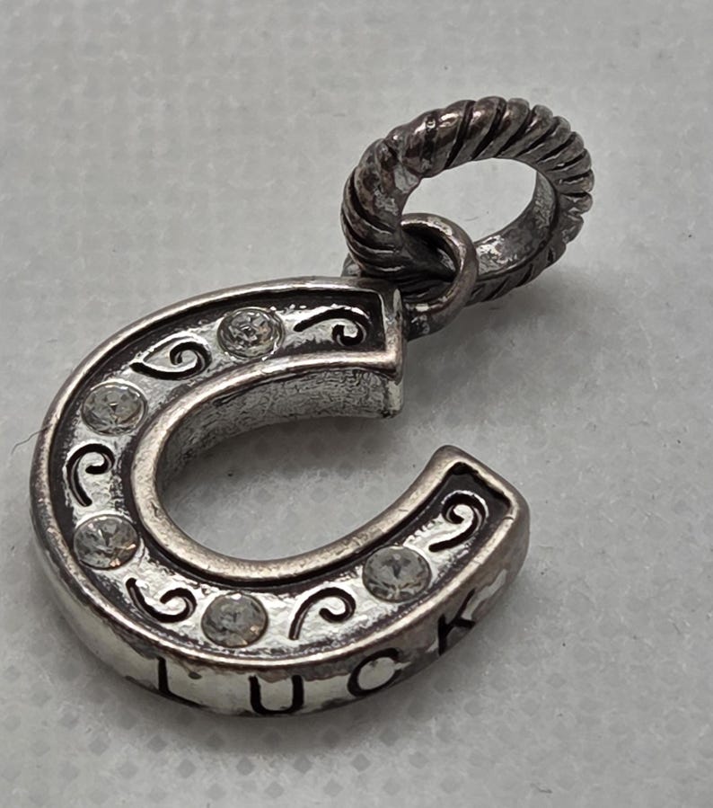 May include: Silver horseshoe charm pendant with clear crystal accents and the word "LUCK" engraved on the bottom. The horseshoe has decorative swirls and a twisted loop for attaching to a chain or bracelet.
