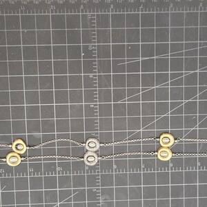 May include: A silver necklace featuring a series of gold and silver circular pendants. The necklace is displayed on a dark grey surface with grid markings. The pendants are spaced along the fine chain.