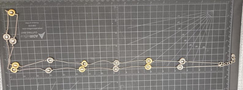 May include: A silver necklace featuring a series of gold and silver circular pendants. The necklace is displayed on a dark grey surface with grid markings. The pendants are spaced along the fine chain.