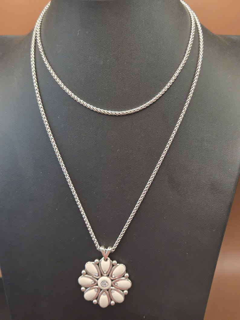 Puede incluir: Un collar plateado con un colgante de flor. El colgante presenta una piedra central transparente rodeada de elementos blancos en forma de p&eacute;talo. El collar tiene un dise&ntilde;o de cadena delicado y se exhibe en un maniqu&iacute; negro.