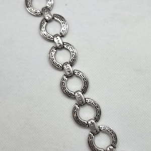 May include: Silver-toned bracelet featuring a series of interconnected, ornate circular links. Each link is detailed with a repeating pattern, creating a vintage or antique aesthetic. The bracelet has a clasp closure.