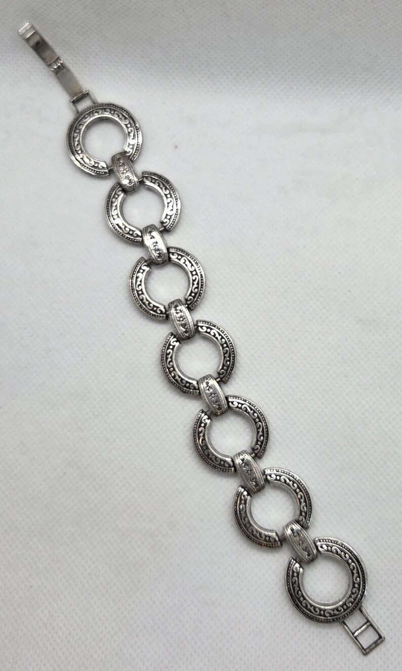 May include: Silver-toned bracelet featuring a series of interconnected, ornate circular links. Each link is detailed with a repeating pattern, creating a vintage or antique aesthetic. The bracelet has a clasp closure.
