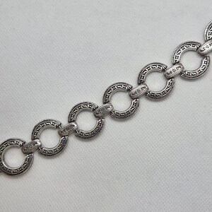 May include: Silver-toned bracelet featuring a series of interconnected, circular links. Each link is adorned with an intricate, embossed pattern. The bracelet has a clasp closure for secure wear. A decorative jewelry piece.