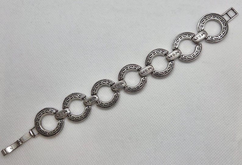 May include: Silver-toned bracelet featuring a series of interconnected, circular links. Each link is adorned with an intricate, embossed pattern. The bracelet has a clasp closure for secure wear. A decorative jewelry piece.