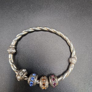 Puede incluir: Brazalete plateado con dise&ntilde;o trenzado. El brazalete presenta cuatro cuentas decorativas con pedrer&iacute;a azul, naranja y rosa. Pedrer&iacute;a transparente adicional adorna los extremos del brazalete.