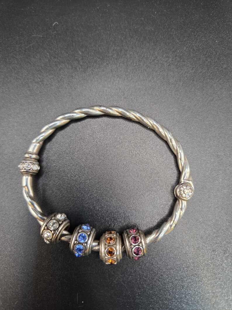 Puede incluir: Brazalete plateado con dise&ntilde;o trenzado. El brazalete presenta cuatro cuentas decorativas con pedrer&iacute;a azul, naranja y rosa. Pedrer&iacute;a transparente adicional adorna los extremos del brazalete.