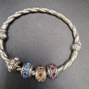 Puede incluir: Pulsera plateada con un dise&ntilde;o de banda trenzada. La pulsera presenta varias cuentas decorativas con piedras preciosas claras, azules, naranjas y rosas. La pulsera tiene un cierre con piedras preciosas claras y un dise&ntilde;o floral.