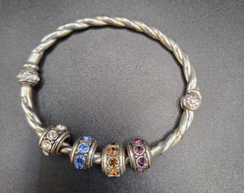 Puede incluir: Pulsera plateada con un dise&ntilde;o de banda trenzada. La pulsera presenta varias cuentas decorativas con piedras preciosas claras, azules, naranjas y rosas. La pulsera tiene un cierre con piedras preciosas claras y un dise&ntilde;o floral.