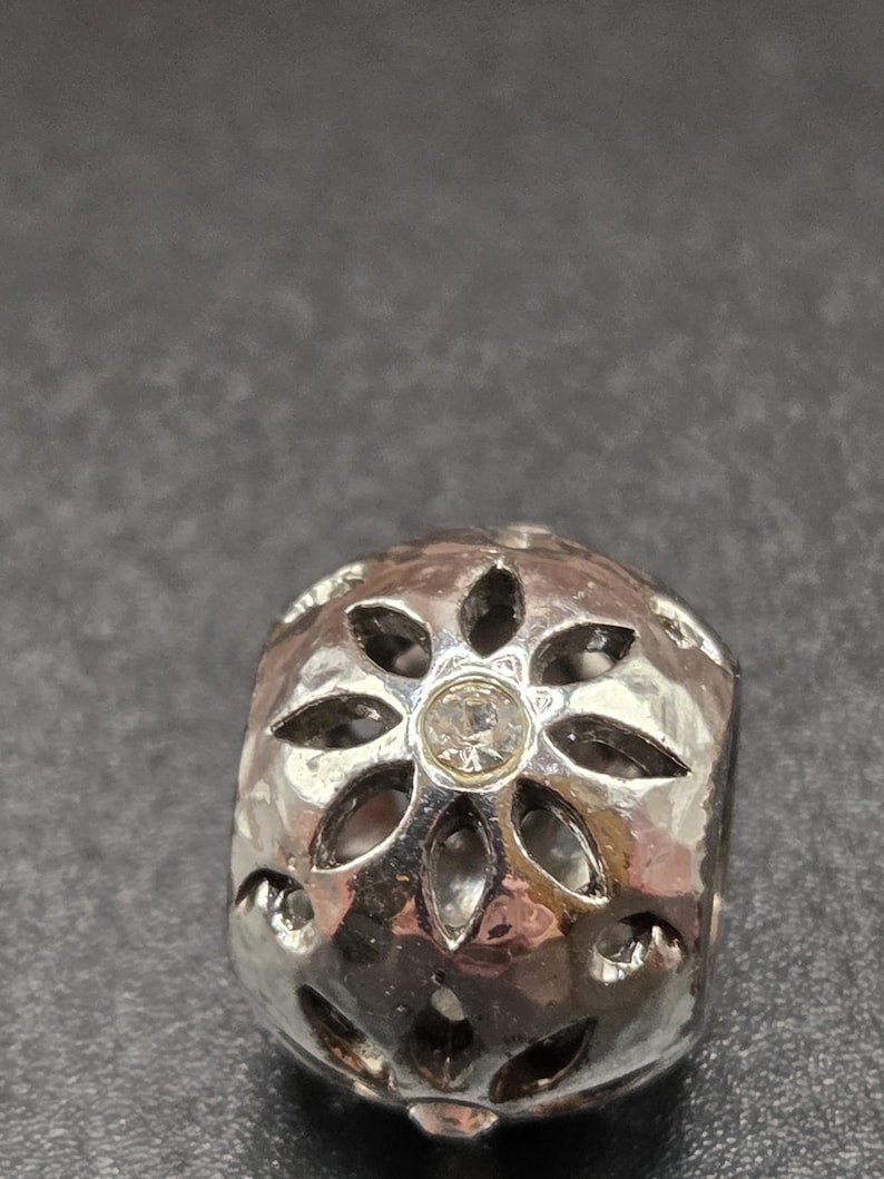 May include: A silver-colored bead with a floral design. The bead has a hammered texture and a small, clear gemstone in the center. The floral design is made up of six petals, each with a pointed end. The bead is cylindrical in shape.