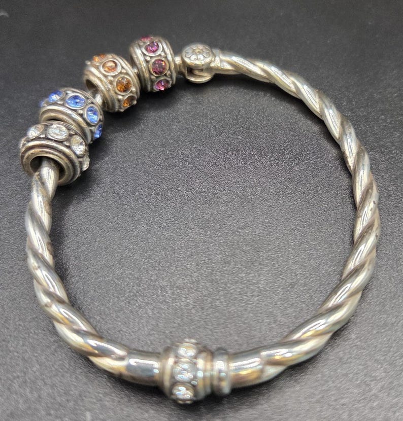 Puede incluir: Pulsera plateada con dise&ntilde;o retorcido. La pulsera presenta cuentas decorativas con piedras preciosas de colores azul, naranja y rosa. Es una pieza de joyer&iacute;a.