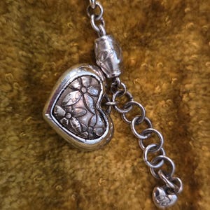 May include: A silver-toned necklace with a heart-shaped pendant. The pendant features a floral design and is connected to a chain with a small heart charm at the end. The necklace is displayed against a textured, golden-brown background.