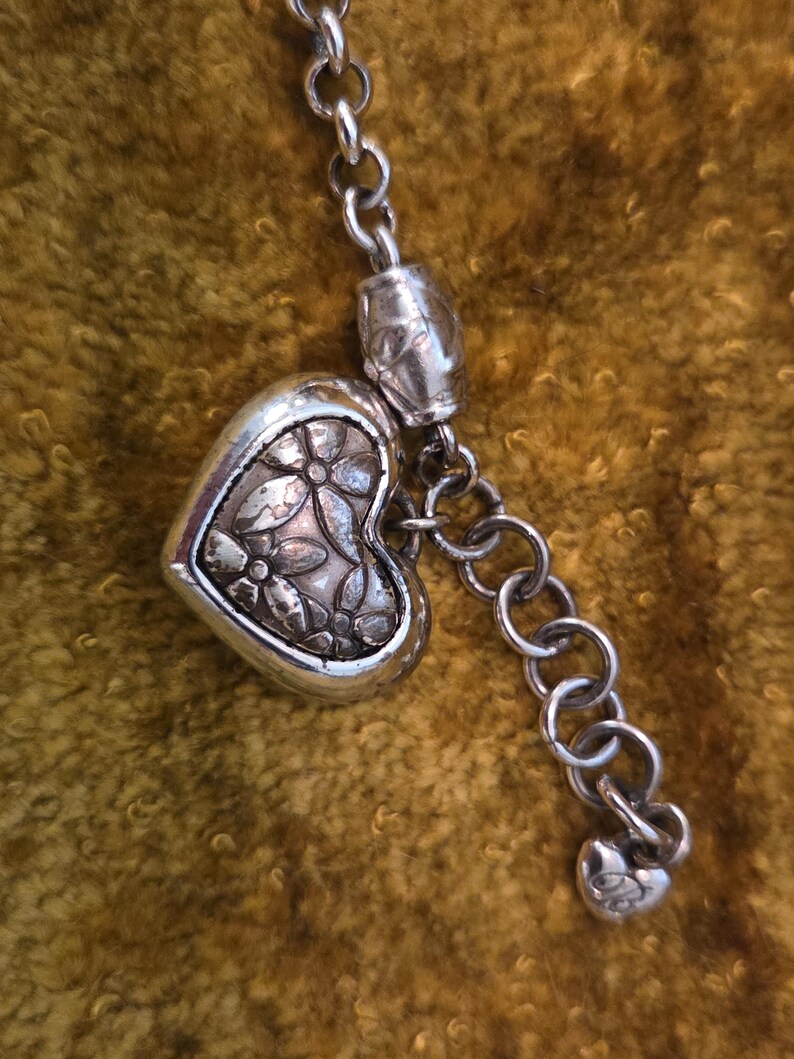 May include: A silver-toned necklace with a heart-shaped pendant. The pendant features a floral design and is connected to a chain with a small heart charm at the end. The necklace is displayed against a textured, golden-brown background.