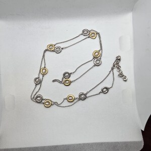 May include: A silver necklace with gold-coloured detailing. The necklace has a fine chain with evenly spaced circular pendants. The pendants have a silver outer ring and a gold-coloured centre. The necklace is displayed on a white surface.