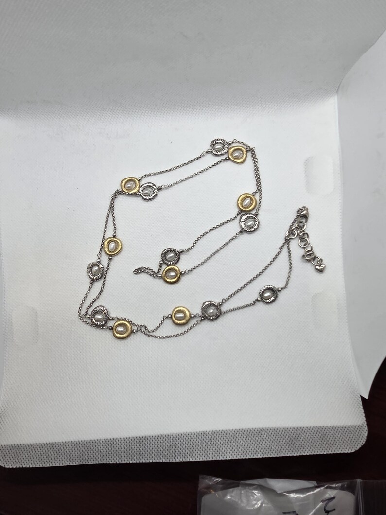 May include: A silver necklace with gold-coloured detailing. The necklace has a fine chain with evenly spaced circular pendants. The pendants have a silver outer ring and a gold-coloured centre. The necklace is displayed on a white surface.