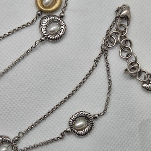 May include: Silver necklace with multiple strands and decorative elements. The necklace features a gold-toned pendant with a pearl, and other pendants with pearls set in ornate silver frames. The chain has a lobster clasp.