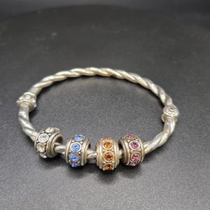 Puede incluir: Pulsera plateada con dise&ntilde;o retorcido. La pulsera presenta cuatro cuentas decorativas con piedras preciosas transparentes, azules, naranjas y rosas. Es una joya.