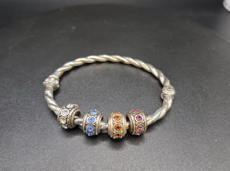 Puede incluir: Pulsera plateada con dise&ntilde;o retorcido. La pulsera presenta cuatro cuentas decorativas con piedras preciosas transparentes, azules, naranjas y rosas. Es una joya.