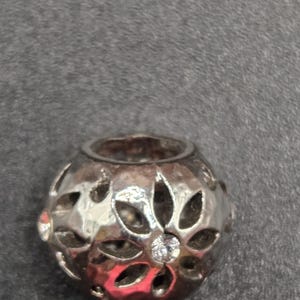 May include: A silver-colored bead with a floral design. The bead has cut-out petal shapes and a small, clear gemstone in the center. The bead is round and has a hole running through the center.