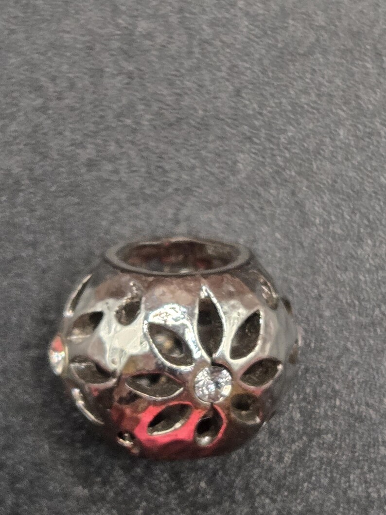 May include: A silver-colored bead with a floral design. The bead has cut-out petal shapes and a small, clear gemstone in the center. The bead is round and has a hole running through the center.