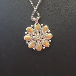 Puede incluir: Un collar plateado con un colgante en forma de flor. El colgante presenta p&eacute;talos dorados, peque&ntilde;as piedras transparentes y una piedra central transparente. La cadena es de dise&ntilde;o tejido. El collar se muestra sobre un fondo negro.