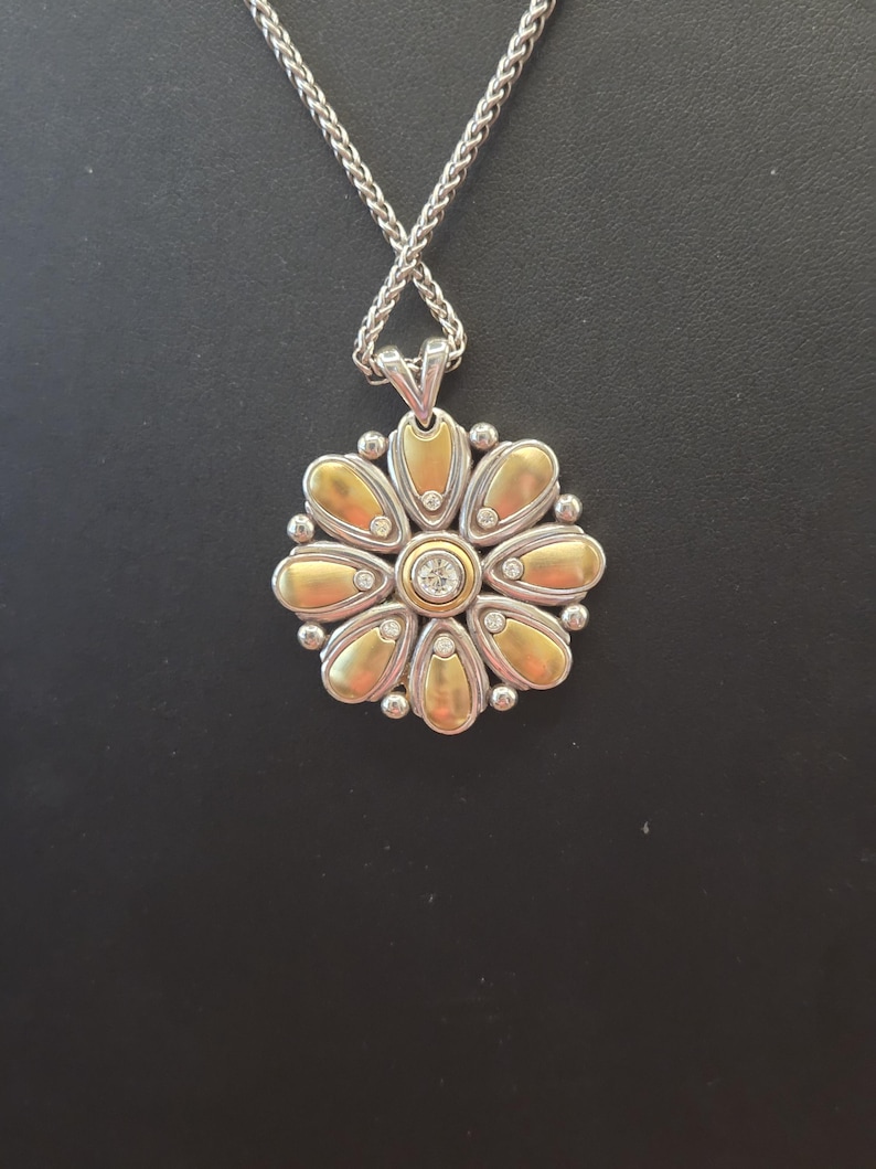 Puede incluir: Un collar plateado con un colgante en forma de flor. El colgante presenta p&eacute;talos dorados, peque&ntilde;as piedras transparentes y una piedra central transparente. La cadena es de dise&ntilde;o tejido. El collar se muestra sobre un fondo negro.