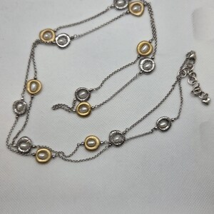 May include: A silver-toned necklace with gold-coloured accents and faux pearls. The necklace features a delicate chain with circular pendants. The pendants alternate between gold and silver, each with a small pearl in the centre. The necklace has an adjustable clasp.