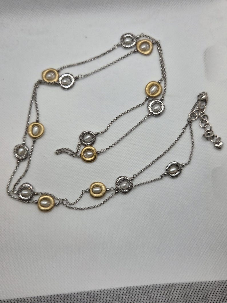 May include: A silver-toned necklace with gold-coloured accents and faux pearls. The necklace features a delicate chain with circular pendants. The pendants alternate between gold and silver, each with a small pearl in the centre. The necklace has an adjustable clasp.