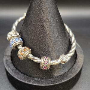 Puede incluir: Pulsera plateada con dise&ntilde;o retorcido. La pulsera presenta varias cuentas decorativas con gemas coloridas y brillantes en tonos azules, naranjas y rosas. La pulsera se exhibe sobre un soporte de terciopelo negro.
