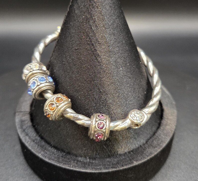 Puede incluir: Pulsera plateada con dise&ntilde;o retorcido. La pulsera presenta varias cuentas decorativas con gemas coloridas y brillantes en tonos azules, naranjas y rosas. La pulsera se exhibe sobre un soporte de terciopelo negro.