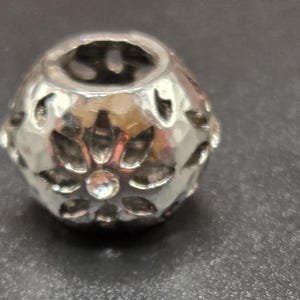 May include: A silver-colored bead with a floral design and a faceted surface. The bead has a central hole and is adorned with small, clear crystals. The bead is likely used for jewelry making or crafting.